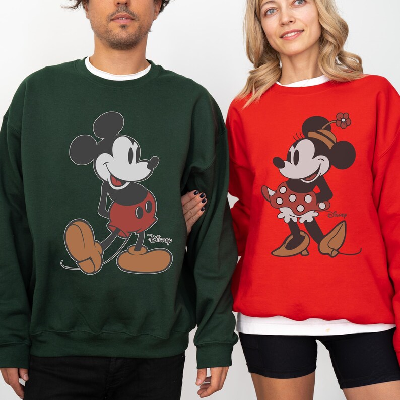 May include: Two sweatshirts featuring Mickey and Minnie Mouse. One is dark green with Mickey Mouse in red shorts and brown shoes. The other is red with Minnie Mouse in a polka dot skirt and brown shoes. Both have the Disney logo.