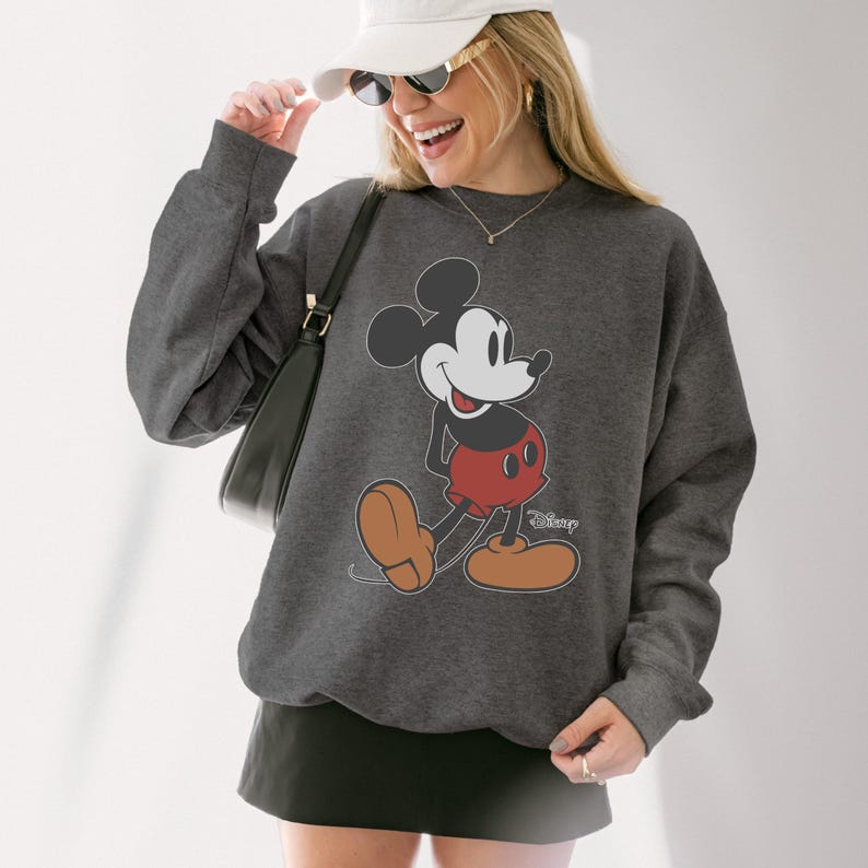 May include: A dark gray sweatshirt featuring a large graphic of Mickey Mouse in his classic pose. Mickey is depicted in black, red, and tan colors. The Disney logo is visible on the lower right side of the graphic.