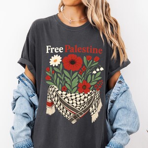 May include: Dark gray t-shirt with the text "Free Palestine" in white and red. The shirt features a floral design with red poppies, white flowers, and green leaves, along with a black and white keffiyeh scarf.
