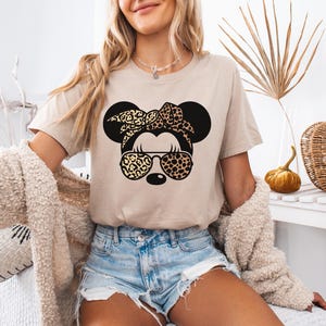 May include: Beige t-shirt featuring a black graphic of a mouse head with a leopard print bow and sunglasses. The mouse head has large black ears. The shirt is paired with denim shorts. The overall theme is casual and fun.