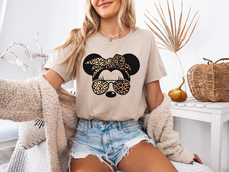 May include: Beige t-shirt featuring a black graphic of a mouse head with a leopard print bow and sunglasses. The mouse head has large black ears. The shirt is paired with denim shorts. The overall theme is casual and fun.