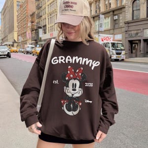 K&ouml;nnte beinhalten: Ein braunes Sweatshirt mit einem Minnie Mouse-Motiv, einer roten Schleife und dem Wort "Grammy" in Wei&szlig;. Das Sweatshirt hat auch den Text "est. 2026" und das Disney-Logo. Die Person auf dem Bild tr&auml;gt eine beige Baseballkappe.