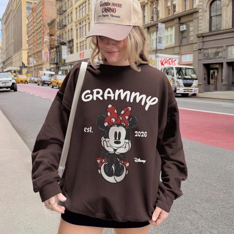 K&ouml;nnte beinhalten: Ein braunes Sweatshirt mit einem Minnie Mouse-Motiv, einer roten Schleife und dem Wort "Grammy" in Wei&szlig;. Das Sweatshirt hat auch den Text "est. 2026" und das Disney-Logo. Die Person auf dem Bild tr&auml;gt eine beige Baseballkappe.