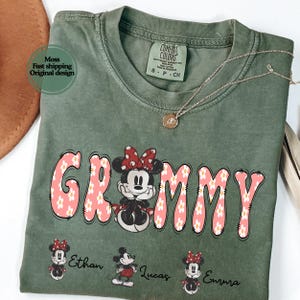 May include: Green t-shirt with the word "Grammy" in pink letters with white daisy accents and a Minnie Mouse graphic. Below the word are three small Minnie Mouse graphics with the names Ethan, Lucas, and Emma.