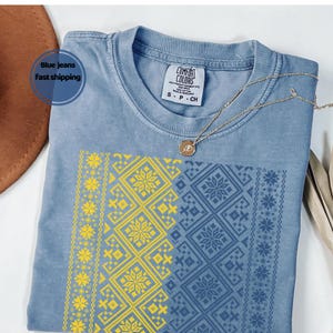May include: A blue Comfort Colors t-shirt with a gold necklace. The shirt features a geometric design in yellow and blue. The text "Blue jeans fast shipping" is in a blue circle.