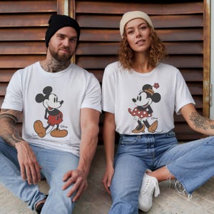 May include: White t-shirts featuring Mickey and Minnie Mouse. Mickey is depicted in his classic outfit, while Minnie wears a polka-dot dress and a flower. The shirts are worn by two people, sitting in front of a brown, weathered wall.