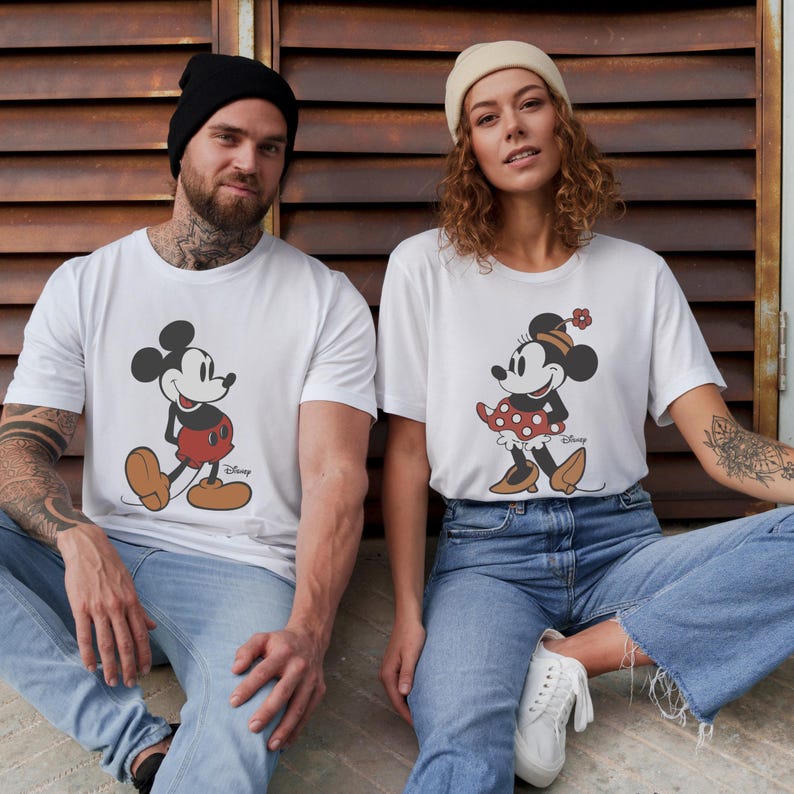May include: White t-shirts featuring Mickey and Minnie Mouse. Mickey is depicted in his classic outfit, while Minnie wears a polka-dot dress and a flower. The shirts are worn by two people, sitting in front of a brown, weathered wall.