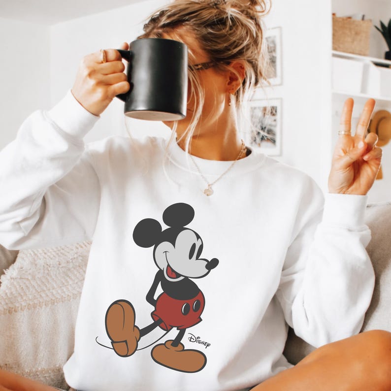 May include: White sweatshirt featuring a classic Mickey Mouse graphic. The cartoon character is depicted in his iconic red shorts, brown shoes, and black and white features. The Disney logo is printed below the character.