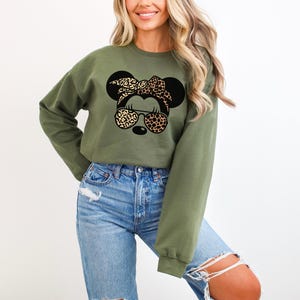 May include: Olive green sweatshirt featuring a graphic of a mouse with a leopard print bow, sunglasses, and ears. The sweatshirt has long sleeves and a crew neck. The mouse graphic is black and tan.