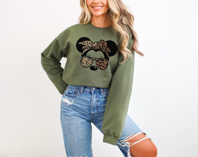 May include: Olive green sweatshirt featuring a graphic of a mouse with a leopard print bow, sunglasses, and ears. The sweatshirt has long sleeves and a crew neck. The mouse graphic is black and tan.