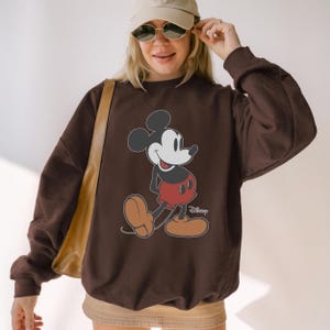 May include: A brown sweatshirt featuring a classic Mickey Mouse graphic. The cartoon character is depicted in his signature red shorts, brown shoes, and black ears. The Disney logo is visible on the bottom right.
