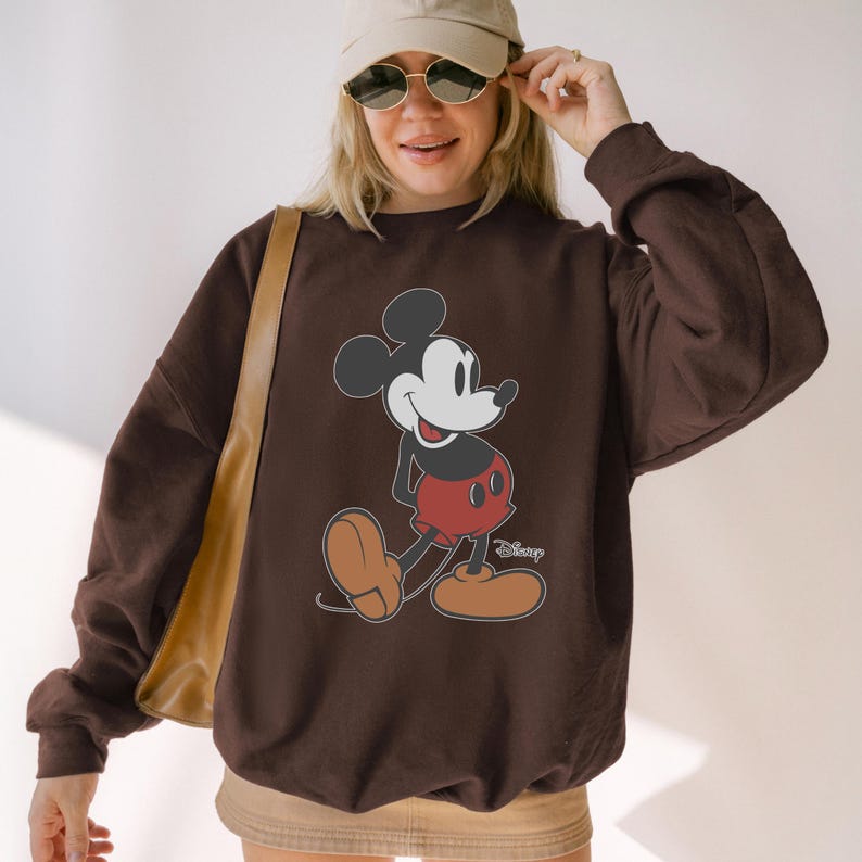 May include: A brown sweatshirt featuring a classic Mickey Mouse graphic. The cartoon character is depicted in his signature red shorts, brown shoes, and black ears. The Disney logo is visible on the bottom right.