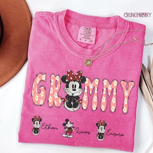 May include: A pink t-shirt with the word "Grammy" in a floral design, featuring Minnie Mouse. The shirt also has the names "Ethan", "Lucas", and "Emma" printed below. A gold necklace and a brown hat are visible.