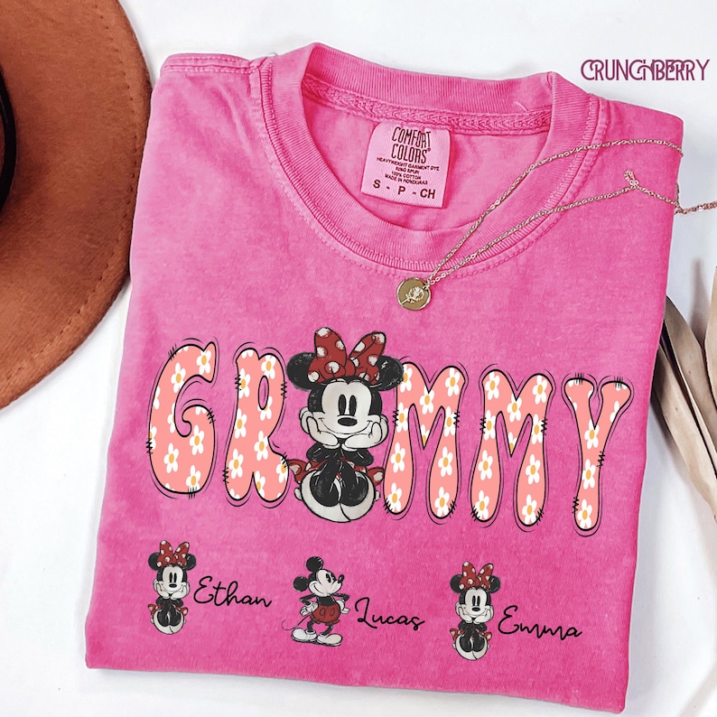 May include: A pink t-shirt with the word "Grammy" in a floral design, featuring Minnie Mouse. The shirt also has the names "Ethan", "Lucas", and "Emma" printed below. A gold necklace and a brown hat are visible.