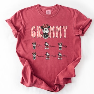 May include: A red t-shirt with the word "Grammy" in a floral design, featuring a Minnie Mouse graphic. Below are smaller Minnie Mouse graphics with names: Ethan, Lucas, Lily, Emma, Noah, and Alex. The shirt has a crew neck and short sleeves.