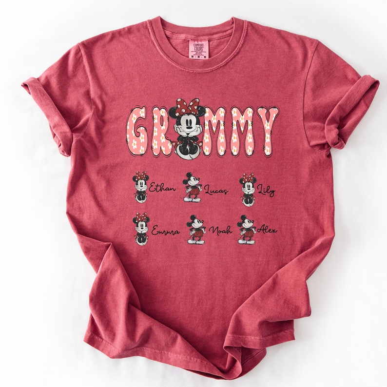 May include: A red t-shirt with the word "Grammy" in a floral design, featuring a Minnie Mouse graphic. Below are smaller Minnie Mouse graphics with names: Ethan, Lucas, Lily, Emma, Noah, and Alex. The shirt has a crew neck and short sleeves.