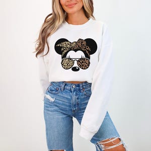 May include: White sweatshirt featuring a graphic of a cartoon mouse with a leopard print bow, sunglasses, and ears. The mouse graphic is in black and tan. The sweatshirt has long sleeves and a crew neck.