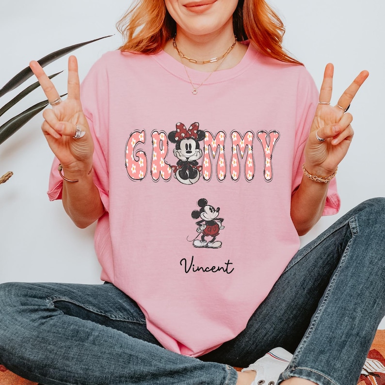 May include: Pink t-shirt featuring the word "Grammy" in a floral design, with a Minnie Mouse graphic. Below, a smaller Mickey Mouse graphic and the name "Vincent" in script. The shirt is a light pink color.