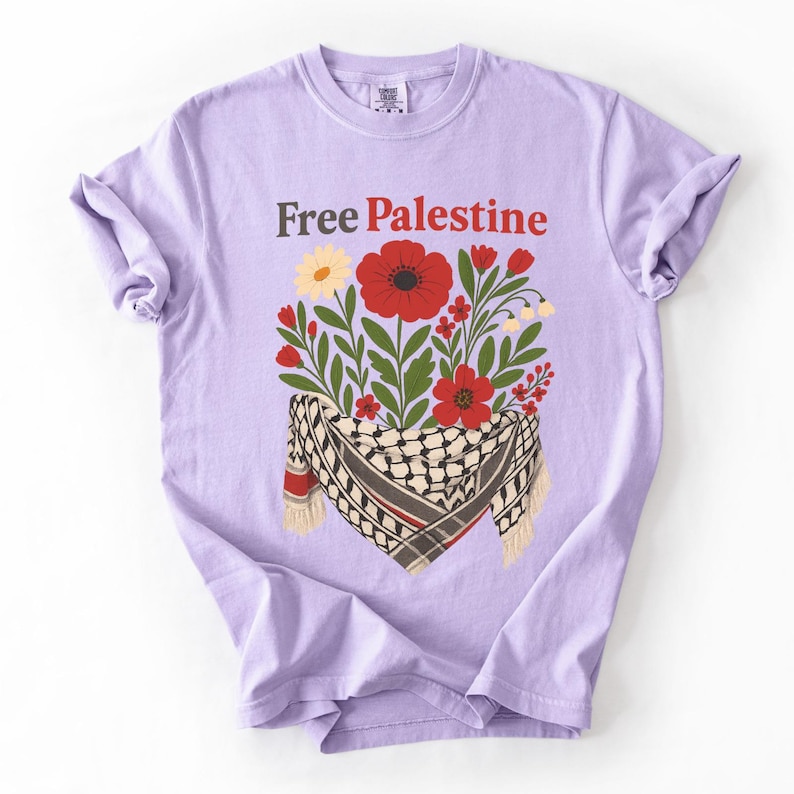 May include: A lilac-colored t-shirt with the text "Free Palestine" in brown and red. The shirt features a floral design with red poppies, yellow flowers, and a black and white scarf.