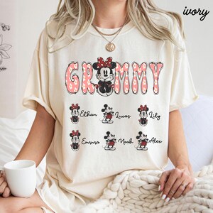 May include: Ivory-colored t-shirt with the word "Grammy" in pink, floral-filled letters. A Minnie Mouse graphic is in the center. Below are Minnie Mouse graphics with names: Ethan, Lucas, Lily, Emma, Noah, and Alex.