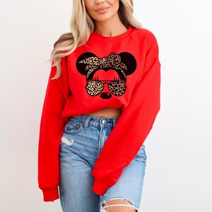 May include: A red sweatshirt featuring a Minnie Mouse graphic with a leopard print bow and sunglasses. The graphic is black and tan. The model is wearing blue jeans.