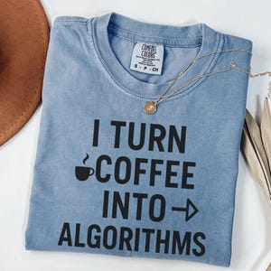 Puede incluir: Una camiseta azul claro con el texto "I turn coffee into algorithms" en negro. Un pequeño icono de taza de café está por encima de la palabra "coffee".