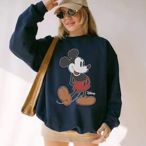 May include: Navy blue sweatshirt featuring a classic Mickey Mouse graphic. The cartoon character is depicted in his signature red shorts, brown shoes, and white gloves. The Disney logo is visible on the bottom right.