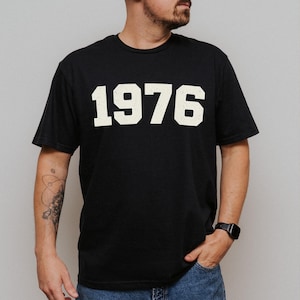 Könnte beinhalten: Schwarzes T-Shirt mit der Jahreszahl "1976" in einer kräftigen, cremefarbenen Schrift auf der Brust. Das Shirt hat kurze Ärmel und einen klassischen Rundhalsausschnitt. Die Person trägt blaue Jeans und eine schwarze Uhr.