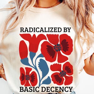 Peut inclure: T-shirt crème avec un motif floral rouge et bleu, et le texte "RADICALIZED BY BASIC DECENCY" en noir. Le motif comprend des fleurs et des feuilles stylisées.