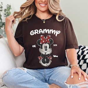 K&ouml;nnte beinhalten: Ein braunes T-Shirt mit dem Wort "Grammy" in wei&szlig;er Schrift &uuml;ber einer Minnie Mouse-Grafik mit roter gepunkteter Schleife. Das Shirt enth&auml;lt auch "est. 2026" und das Disney-Logo.
