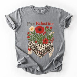 May include: Gray t-shirt with the text "Free Palestine" in brown and red. The shirt features a floral design with red poppies, white flowers, and a keffiyeh scarf.