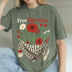 May include: Olive green t-shirt with the text "Free Palestine" above a floral design. The design includes red poppies, white flowers, and a black and white scarf with red accents.