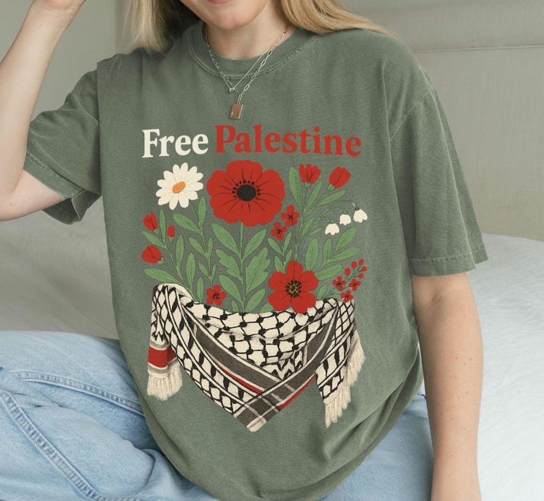 May include: Olive green t-shirt with the text "Free Palestine" above a floral design. The design includes red poppies, white flowers, and a black and white scarf with red accents.