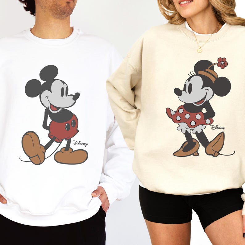 May include: White and beige sweatshirts featuring Mickey and Minnie Mouse. Mickey is in red shorts and brown shoes, while Minnie wears a red polka-dot dress and brown shoes. Both sweatshirts have the Disney logo.