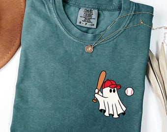 May include: Teal Comfort Colors t-shirt with a baseball-themed ghost graphic. The ghost wears a red baseball cap and holds a bat. A gold necklace with a pendant is draped on the shirt. The shirt has the Comfort Colors label.