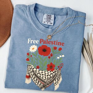 May include: A light blue t-shirt with the words "Free Palestine" in red. The shirt features a floral design with red poppies, white daisies, and other flowers, along with a black and white scarf design.