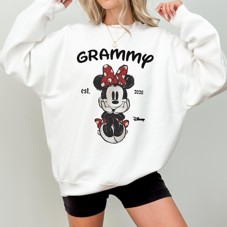 K&ouml;nnte beinhalten: Wei&szlig;es Sweatshirt mit dem Wort "Grammy" in schwarzer Schrift &uuml;ber einer Minnie Mouse-Grafik. Die Grafik enth&auml;lt eine rote Schleife mit wei&szlig;en Punkten, den Text "est. 2026" und das Disney-Logo.