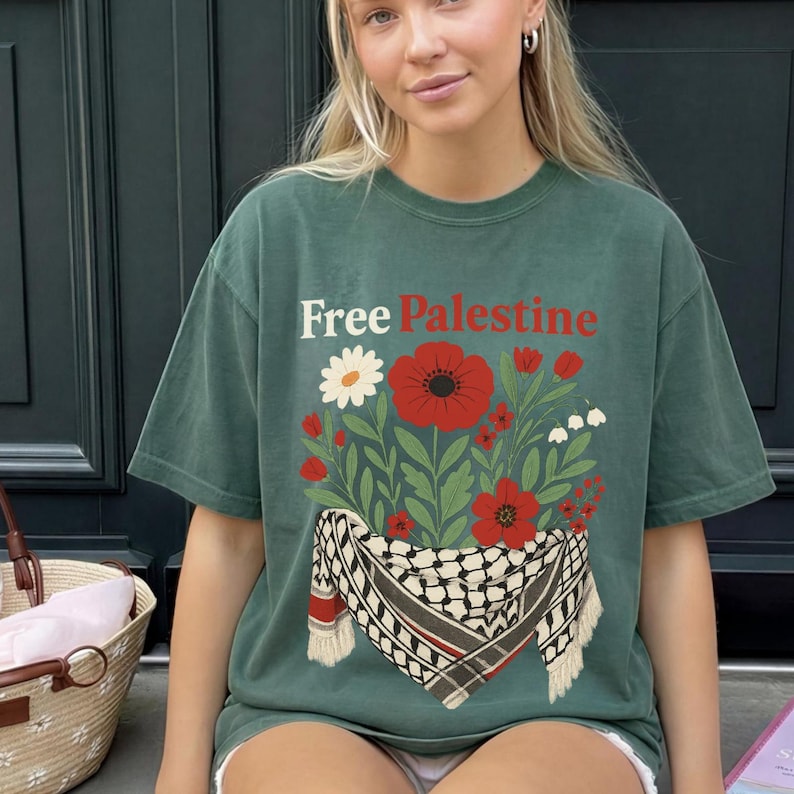 May include: Green t-shirt with the text "Free Palestine" in white and red. The shirt features a floral design with red poppies, white daisies, and a traditional Palestinian scarf.