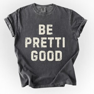 May include: Dark gray t-shirt with the phrase "BE PRETTI GOOD" printed in a distressed, off-white font. The shirt has rolled-up sleeves and a crew neck. The fabric appears soft and worn.