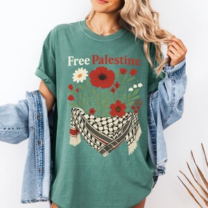 May include: Green t-shirt with the text "Free Palestine" in white and red. The shirt features a floral design with red poppies, white flowers, and a black and white scarf. The shirt has short sleeves.