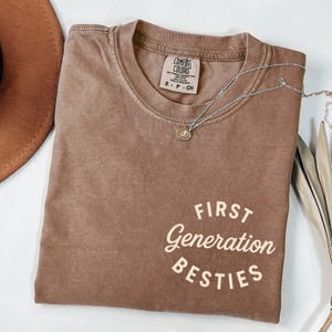 May include: A brown t-shirt with the text "FIRST GENERATION BESTIES" in a cream colour. The shirt is made of a soft, comfortable material and has a relaxed fit.