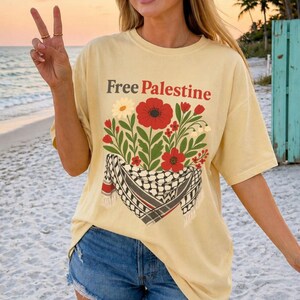 May include: A light yellow t-shirt with the text "Free Palestine" in brown. The shirt features a floral design with red poppies, white daisies, and green leaves, along with a black and white checkered scarf.