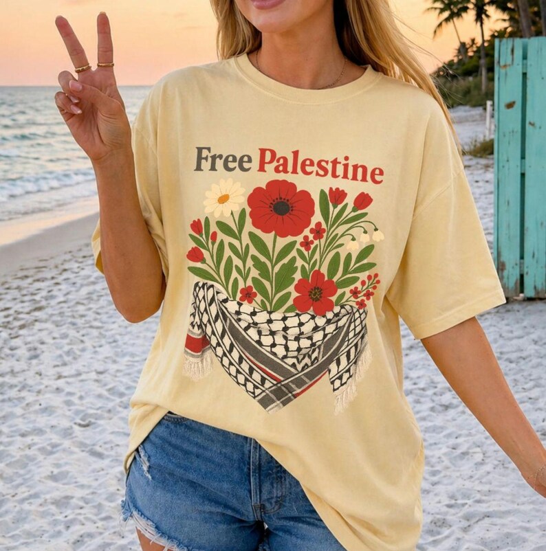 May include: A light yellow t-shirt with the text "Free Palestine" in brown. The shirt features a floral design with red poppies, white daisies, and green leaves, along with a black and white checkered scarf.