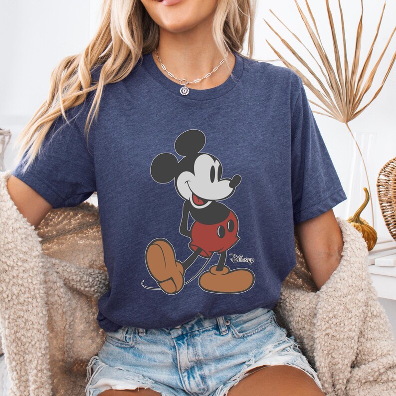 May include: Navy blue t-shirt featuring a classic Mickey Mouse graphic. The cartoon character is depicted in his iconic red shorts, black ears, and brown shoes. The Disney logo is visible below the character.