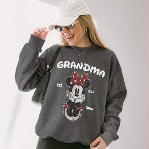 Op de afbeelding: Een donkergrijze sweatshirt met het woord "GRANDMA" in witte letters. Daaronder een Minnie Mouse-afbeelding met een rode strik. De sweatshirt heeft ook de tekst "est. 2024" en het Disney-logo. Een casual, comfortabel kledingstuk.