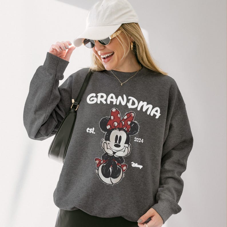 Op de afbeelding: Een donkergrijze sweatshirt met het woord "GRANDMA" in witte letters. Daaronder een Minnie Mouse-afbeelding met een rode strik. De sweatshirt heeft ook de tekst "est. 2024" en het Disney-logo. Een casual, comfortabel kledingstuk.