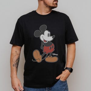 May include: Black t-shirt featuring a classic Mickey Mouse graphic. The cartoon character is depicted in his iconic pose, with red shorts, brown shoes, and white gloves. The Disney logo is visible.