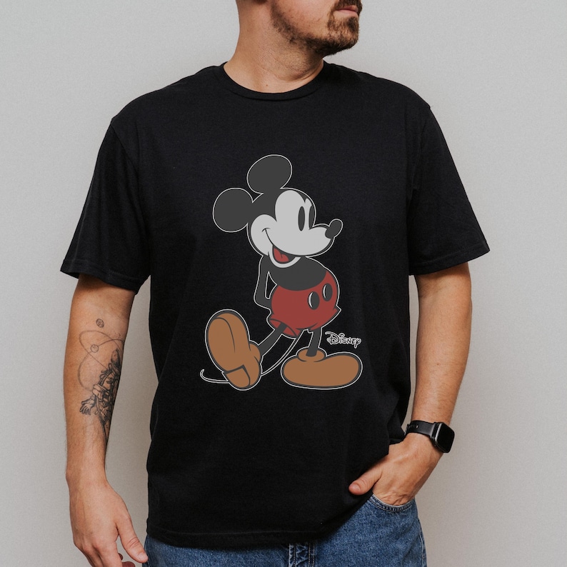 May include: Black t-shirt featuring a classic Mickey Mouse graphic. The cartoon character is depicted in his iconic pose, with red shorts, brown shoes, and white gloves. The Disney logo is visible.