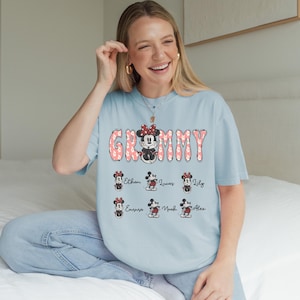 May include: Light blue t-shirt with the word "Grammy" in pink floral letters and a Minnie Mouse graphic. Below the word are six Minnie Mouse graphics with names: Ethan, Lucas, Lily, Emma, Noah, and Alex. The shirt is worn by a person.