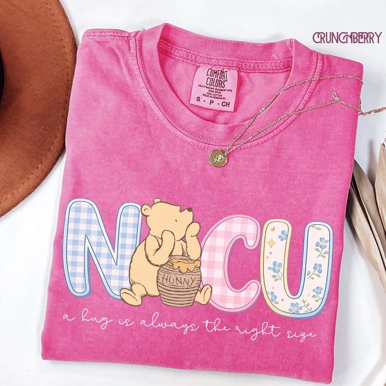 Może przedstawiać: R&oacute;żowa koszulka Comfort Colors z napisem "NICU" w zabawnym kroju pisma. Litery "N" i "C" mają wz&oacute;r w kratkę, a "U" ma kwiatowy wz&oacute;r. Obok siedzi postać z kresk&oacute;wki, niedźwiedź z garnkiem miodu. Poniżej znajduje się napis "a hug is always the right size".